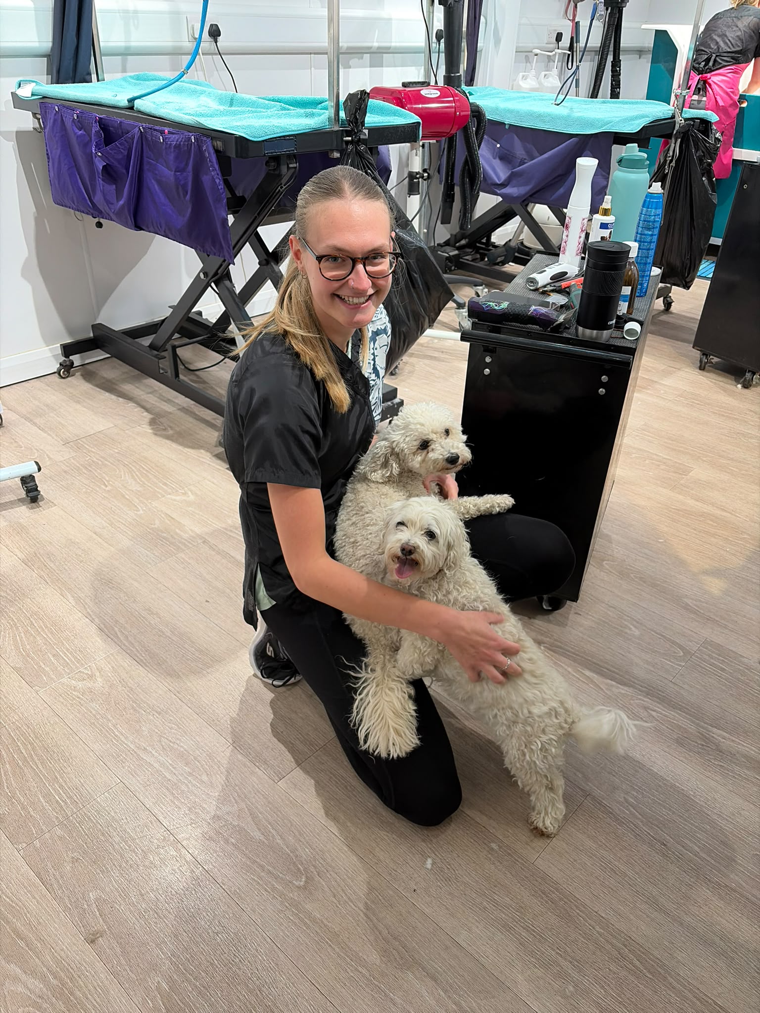 A groomer kneeling on the salon floor while a white doodle rests its paws on her